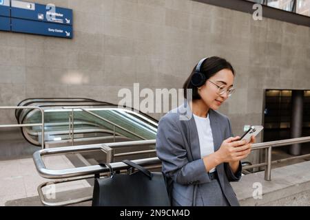 Jeune femme asiatique dans un casque utilisant un smartphone tout en se tenant à l'extérieur près de l'escalier roulant Banque D'Images