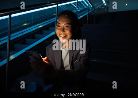 Jeune femme asiatique joyeuse utilisant un téléphone portable tout en étant assise sur les escaliers à côté de l'escalier mécanique Banque D'Images