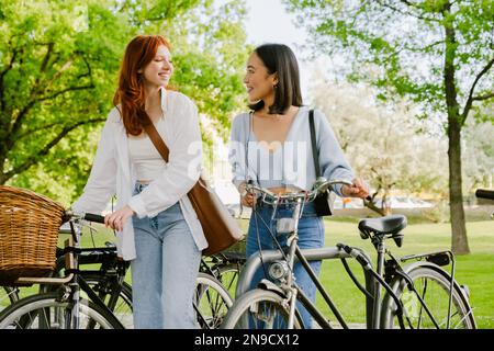 Deux jeunes filles souriantes et souriantes marchant avec des vélos dans le parc et regardant l'une sur l'autre Banque D'Images