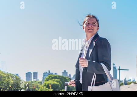 Femme d'affaires tenant un sac et tirant une valise tout en marchant dans la ville. Banque D'Images