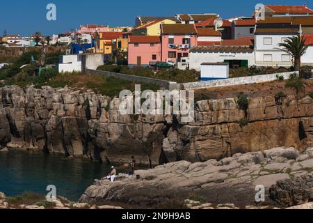 Un couple pêche des rochers au port de Peniche, dans le district de Leiria, sur la côte Atlantique du centre du Portugal, sur fond d’atelier de menuiserie marine et de maisons modernes colorées avec des toits de tuiles rouges et des murs peints en rose, orange, jaune et blanc. Banque D'Images