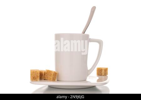 Plusieurs cubes de sucre brun avec une tasse blanche en céramique avec soucoupe et cuillère en métal, macro, isolée sur fond blanc. Banque D'Images