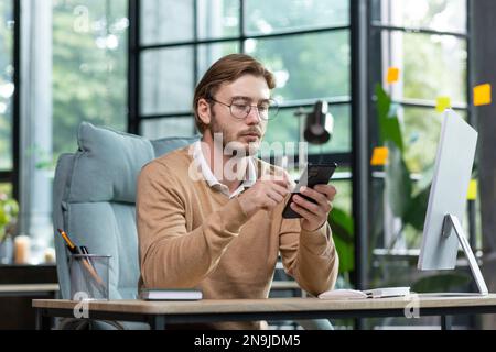 Jeune programmeur sérieux, indépendant assis au bureau, travaillant à l'ordinateur. Tient un téléphone dans ses mains, utilise un gadget. Écrit des messages, vérifie le courrier, discute. Banque D'Images