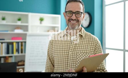 Homme d'âge moyen enseignant souriant confiant tenant la planchette à pince dans la salle de classe Banque D'Images