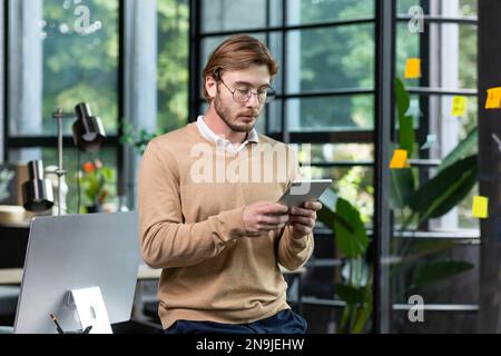 Portrait d'un jeune programmeur masculin qui se tient au bureau à la table et se concentre sur la tablette. Banque D'Images