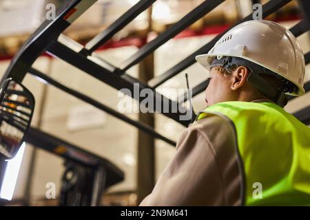 Femme portant un casque de sécurité lors de l'utilisation d'un chariot élévateur dans un entrepôt et parlant à une radio portable Banque D'Images