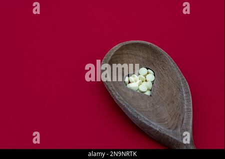 Cuillère en bois avec un trou en forme de coeur rempli d'aspirine à faible dose isolée sur un fond rouge. Concept de santé cardiaque Banque D'Images