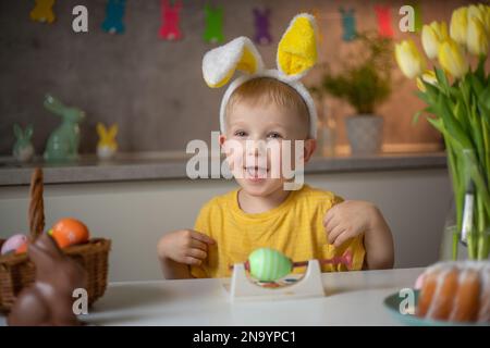 Portrait émotionnel d'un petit garçon gai portant des oreilles de lapin le jour de Pâques qui rit merrement, joue avec des œufs de Pâques colorés assis à une table dedans Banque D'Images