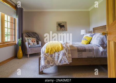 Lit queen size sur cadre en bois brun clair et fauteuil en osier taupe avec coussins de la faune dans la chambre d'amis au sous-sol pièce sur pièce à l'intérieur de la maison. Banque D'Images