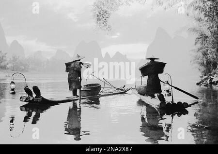 Image en noir et blanc de pêcheurs debout sur des bateaux sur une rivière tranquille dans le brouillard avec des formations karstiques au loin ; Chine Banque D'Images