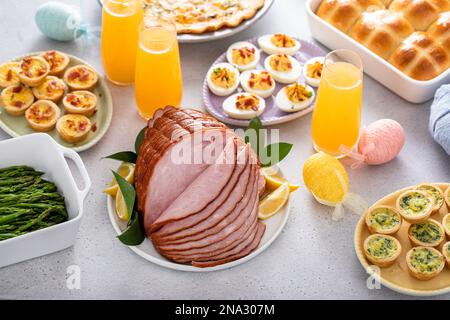 Table de brunch de Pâques avec jambon, quiche, petits pains de croix chaude et œufs dévorés et mimosas Banque D'Images