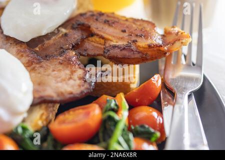 Petit déjeuner copieux : œufs pochés avec bacon sur pain grillé, tomates frites et épinards, jus d'orange, assiette noire avec couteau et fourchette Banque D'Images