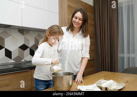 Mère enseignant à sa fille la préparation et le mélange de pâte dans la cuisine intérieure moderne. Peu d'aide. Sourire et s'amuser ensemble. Temps sans g Banque D'Images