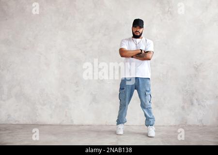 Portrait sur toute la longueur Homme hispanique tient les bras croisés, dans un Jean décontracté et blanc maquette T-shirt et casquette de base-ball, gars cool regarde l'appareil photo contre Banque D'Images