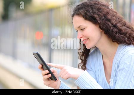 Une femme heureuse vérifie le contenu d'un smartphone assis dans la rue Banque D'Images