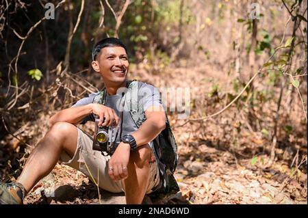 Homme asiatique heureux et actif avec équipement de trekking et son appareil photo rétro se trouve sur le rocher, prenant un repos après la randonnée sur le sentier des montagnes Rocheuses. Banque D'Images