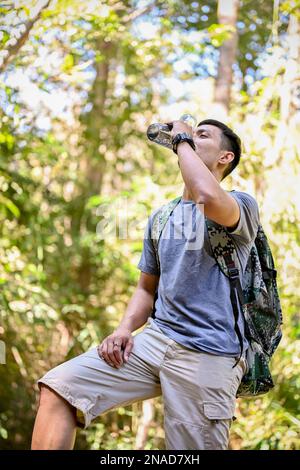 Portrait, fatigué et assoiffé asiatique homme voyageur ou trekker boire l'eau d'une bouteille tout en se reposant sur le sentier de montagne. Activité d'aventure en plein air Banque D'Images