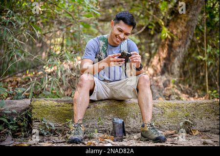 Bon voyageur ou trekker asiatique avec équipement de trekking et sac à dos à l'aide de son smartphone tout en se reposant sur une bûche en bois dans la forêt. Banque D'Images