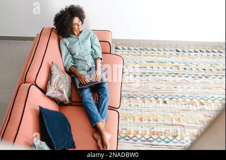Une jeune femme afro-américaine passe ses loisirs à la maison avec un ordinateur portable. Petite fille calme assise dans un canapé confortable et vue sur l'écran de l'ordinateur portable. Navigation sur le Web, visionnage de séries, achats sur le réseau Banque D'Images