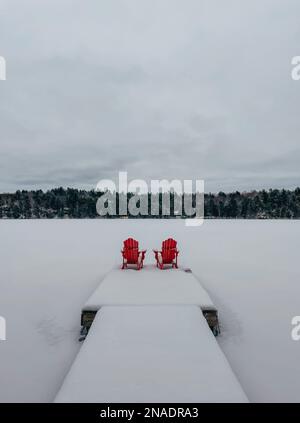 Chaises rouges adirondack au bout d'un quai enneigé sur un lac en hiver. Banque D'Images