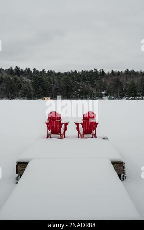 Chaises rouges adirondack au bout d'un quai enneigé sur un lac en hiver. Banque D'Images