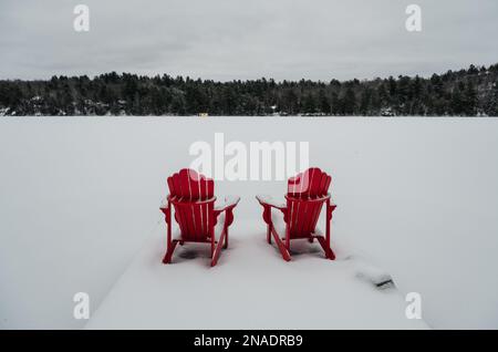 Chaises rouges adirondack au bout d'un quai enneigé sur un lac en hiver. Banque D'Images