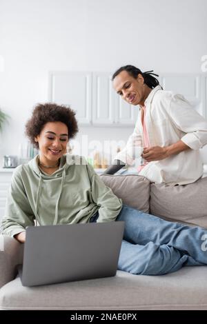 Une femme afro-américaine joyeuse utilisant un ordinateur portable près d'un petit ami avec un café à la maison Banque D'Images