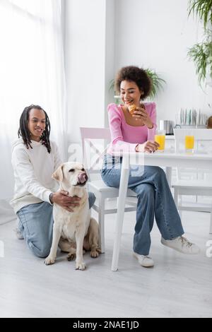 Femme afro-américaine souriante tenant un croissant près d'un petit ami et du labrador dans la cuisine Banque D'Images