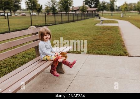 Une jeune fille est assise sur un banc au parc pour chiens, où elle tient des feuilles d'automne Banque D'Images