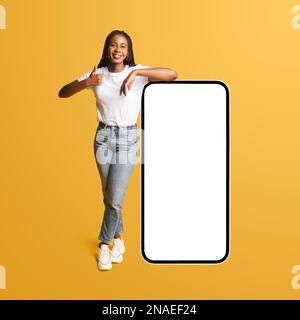 Meilleure affaire, promotion. Souriante femme afro-américaine penchée sur un immense smartphone avec un écran vide, montrant le pouce vers le haut, une étudiante recommandant une nouvelle application ou un nouveau site Web, maquette, isolée Banque D'Images