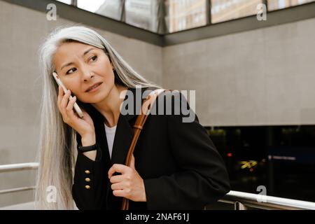 Femme asiatique grise souriant et parlant au téléphone portable en se tenant à l'intérieur Banque D'Images