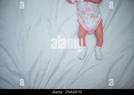 bébé au lit. Nouveau-né en bodie blanche. Pieds de nouveau-né en chaussettes blanches. Vêtements pour enfants pour la croissance. Banque D'Images