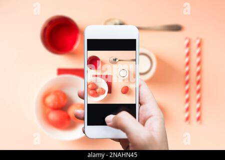 Prenez des photos à la main avec votre smartphone. Œufs de pâques teints en rouge, cuillère, lait, paille et colorant alimentaire sur le plateau de table. Fond rose. Banque D'Images