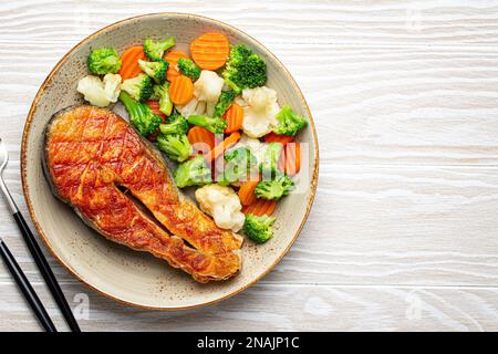 Dîner de poisson sain : steak de saumon grillé avec salade de légumes sur plaque de céramique avec fourchette et couteau sur fond de bois blanc d'en haut avec Banque D'Images