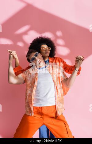 Femme afro-américaine élégante tenant les mains de l'homme brunette dans l'ensemble sur fond rose avec ombre, image de stock Banque D'Images