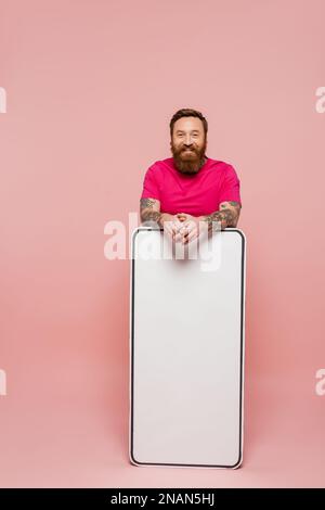 homme barbu et tatoué en t-shirt magenta regardant l'appareil photo près d'un énorme modèle de téléphone sur fond rose, image de stock Banque D'Images