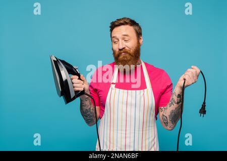 homme barbu sceptique en tablier rayé regardant le fer et souriant isolé sur bleu, image de stock Banque D'Images