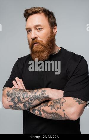 grave homme tatoué dans un t-shirt noir debout avec des bras croisés et regardant la caméra isolée sur gris, image de stock Banque D'Images