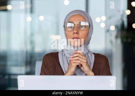 Gros plan. Portrait d'une jeune belle étudiante arabe dans un hijab. Assis à la table, travaillant, étudiant à l'ordinateur. Il regarde soigneusement et sérieusement sur le côté, à la fenêtre. Banque D'Images