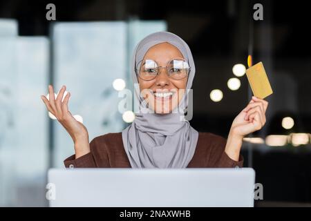 Gros plan. Portrait d'une jeune belle femme arabe dans un hijab assis au bureau à un ordinateur portable, tenant une carte de crédit dans ses mains, souriant à la caméra, posant, faisant de la publicité. Banque D'Images