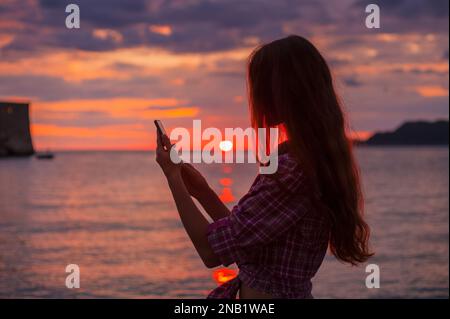 Vue latérale silhouette une jeune femme rousse à l'aide d'un smartphone sur la plage pendant idyllique coucher de soleil coloré mer Adriatique, photo prise Budva Monténégro Banque D'Images