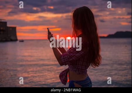 Vue latérale silhouette une jeune femme rousse à l'aide d'un smartphone sur la plage pendant idyllique coucher de soleil coloré mer Adriatique, photo prise Budva Monténégro Banque D'Images