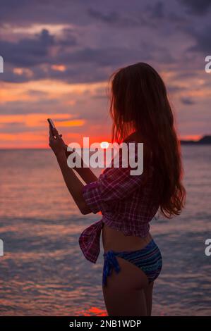 Vue latérale silhouette une jeune femme rousse à l'aide d'un smartphone sur la plage pendant idyllique coucher de soleil coloré mer Adriatique, photo prise Budva Monténégro Banque D'Images