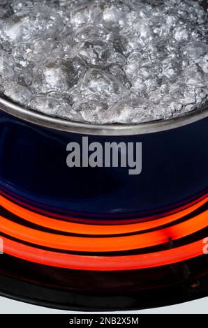 Gros plan vertical d'une casserole bleue sur un dessus de cuisinière sur un brûleur chaud rouge contenant de l'eau bouillante. Seule une partie du pot s'affiche. Banque D'Images