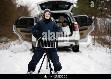 Garçon avec pile de panneau solaire sur trépied contre la voiture dans les bois d'hiver. Banque D'Images