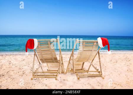 Chaises longues en bois et chapeaux du Père Noël sur la plage près de la mer. Vacances de Noël Banque D'Images