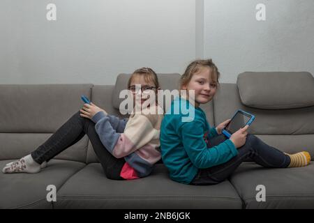 Deux jeunes sœurs s'assoient dos à dos sur le canapé avec leurs tablettes. Ils regardent dans la caméra Banque D'Images