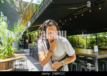 jeune vénézuélien latin avec des dreadlocks, debout à l'extérieur d'un restaurant touchant son menton et regardant avec doute sur le côté Banque D'Images