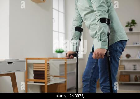 L'homme se penche sur des béquilles, essayant de marcher dans son salon à la maison. Banque D'Images