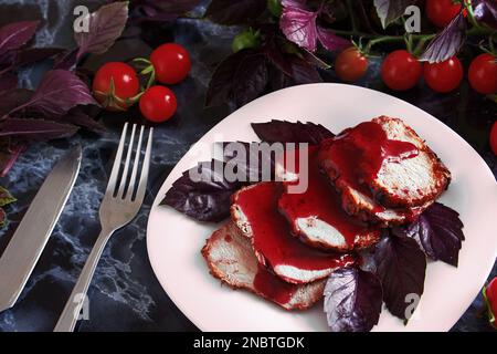 Steak juteux rôti de porc bien cuit avec sauce rouge et feuilles de basilic sur une assiette blanche sur une table en marbre noir. Tomates cerises et brins de basilic sur un mar Banque D'Images